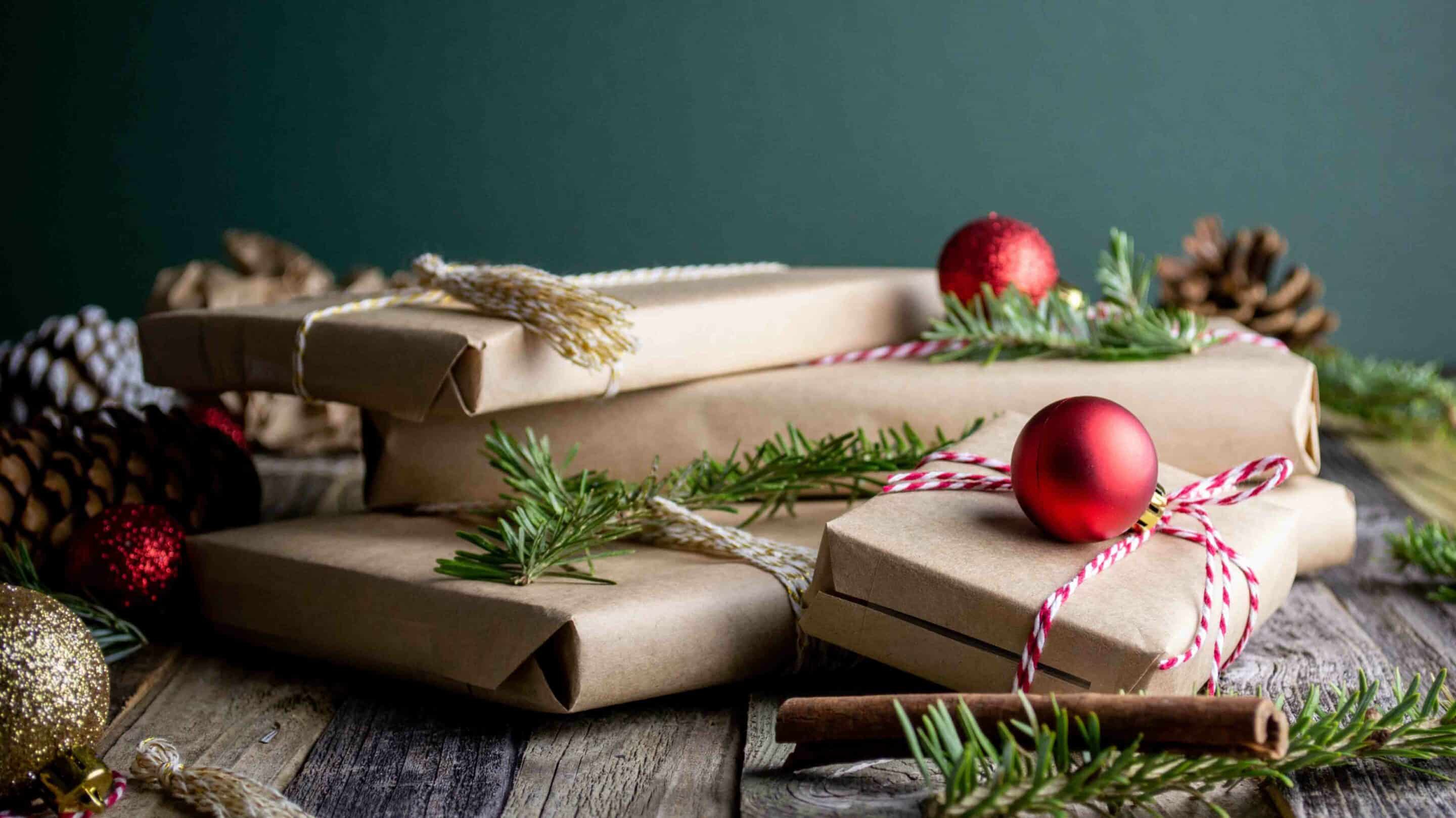 Holiday-themed gifts wrapped in brown paper with red ornaments, pine needles, and pinecones on a wooden surface against a dark green background